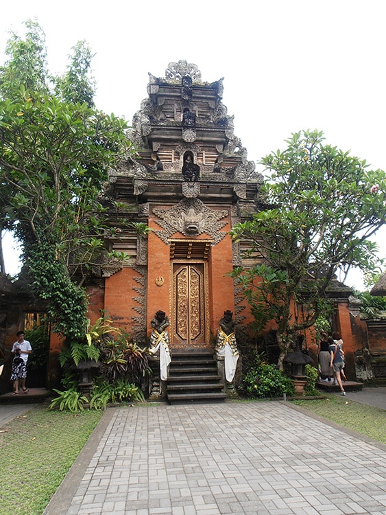 Een prachtig Balinees tempelgebouw met gedetailleerde houtsnijwerken en traditionele architectuur, omringd door groene bomen en planten. Deze serene locatie, perfect voor soloreizen, nodigt uit om de rijke cultuur en spirituele geschiedenis van Bali te ontdekken in alle rust.