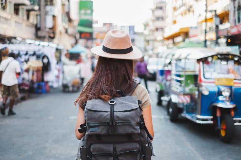 Een vrouwelijke solo-reiziger met een beige hoed en een grote grijze rugzak loopt door een levendige, kleurrijke straat vol tuk-tuks en marktkraampjes—een typisch tafereel dat inspiratie geeft voor leuke dingen om alleen te doen op reis, zoals lokale markten verkennen en cultuur opsnuiven. De sfeer is druk, avontuurlijk en vol ontdekkingen.