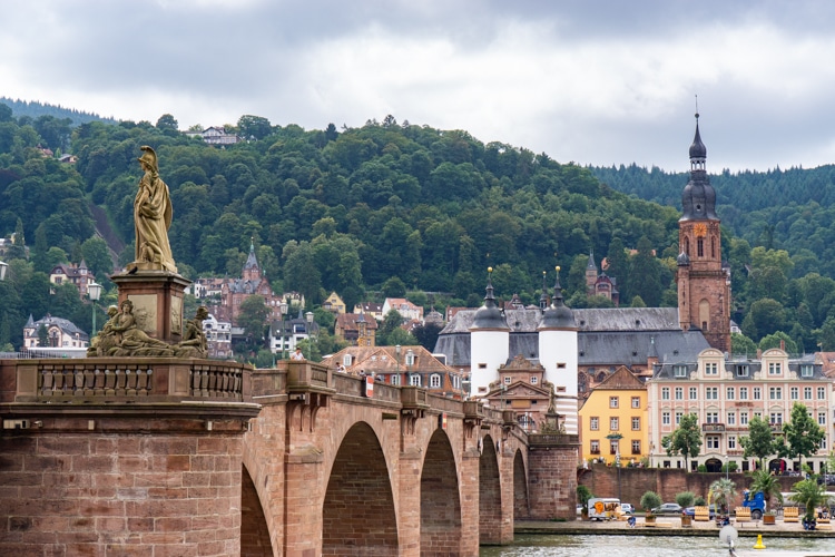 Deze afbeelding toont de historische stad Heidelberg, een van de mooiste steden in Duitsland, met de iconische Alte Brücke (Oude Brug) op de voorgrond. De brug, met een standbeeld en robuuste stenen bogen, leidt naar een sfeervol stadsbeeld met kleurrijke gebouwen, een kerk en beboste heuvels op de achtergrond. Deze charmante bestemming is een perfecte keuze voor liefhebbers van historische steden in Duitsland en schilderachtige uitzichten.