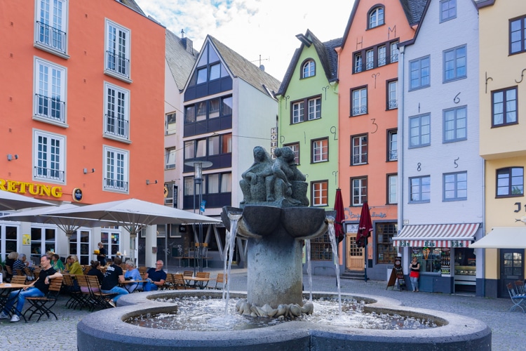 Deze afbeelding toont een sfeervol plein in Keulen, Duitsland, met een fontein op de voorgrond en kleurrijke historische gebouwen op de achtergrond. De gevels zijn versierd met unieke cijfers en letters, wat de straat een speels karakter geeft. Mensen genieten op terrasjes van een ontspannen moment, wat deze plek een leuke plek in Duitsland maakt om te bezoeken.