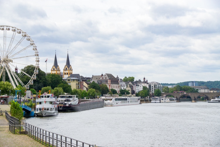 Deze afbeelding toont de schilderachtige stad Koblenz, Duitsland, met een rivier, een groot reuzenrad en historische gebouwen, waaronder een kerk met twee opvallende torens. Langs de kade liggen verschillende boten aangemeerd, wat bijdraagt aan de levendige sfeer van deze plek. Dit is zonder twijfel een leuke plek in Duitsland om te genieten van een ontspannen wandeling langs het water.