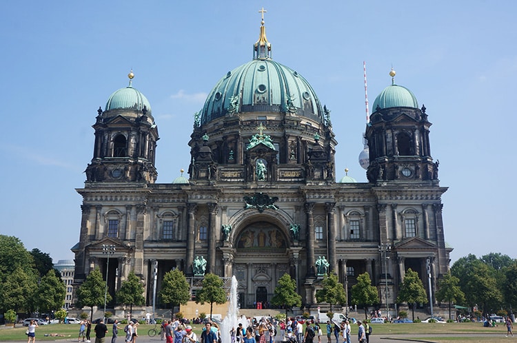 Deze afbeelding toont de Berliner Dom, een prachtige historische kathedraal in Duitsland met een imposante groene koepel en gedetailleerde architectuur. De kathedraal staat tegen een heldere blauwe lucht, met de Fernsehturm (TV-toren) subtiel zichtbaar op de achtergrond. Op de voorgrond wandelen toeristen en bezoekers rond een fontein in een groen park, wat bijdraagt aan de levendige sfeer van deze leuke plek in Duitsland.