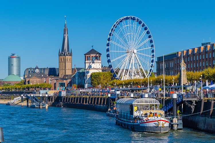Deze afbeelding toont de bruisende Rijnpromenade in Düsseldorf, Duitsland, met een groot reuzenrad, historische gebouwen en een aanlegsteiger met een boot op de voorgrond. De blauwe lucht en het levendige stadsbeeld maken deze locatie extra uitnodigend. Dit is zeker een leuke plek in Duitsland om te genieten van het uitzicht en de gezellige sfeer langs de rivier.