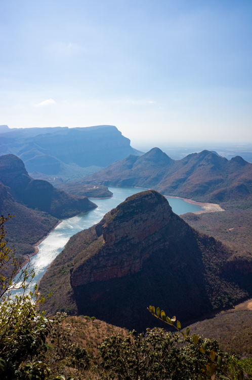 Verticaal uitzicht op de Blyde River Canyon, een van de hoogtepunten van de Panoramaroute in Zuid-Afrika, met een glinsterende rivier die tussen groene kliffen slingert onder een heldere blauwe lucht.

