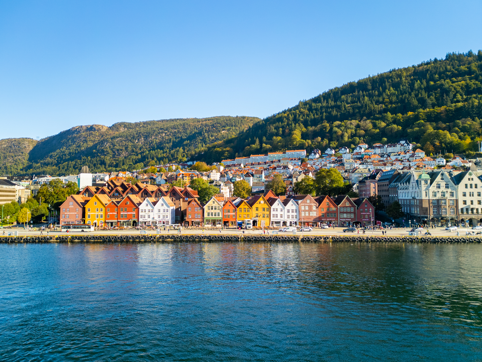 Uitzicht vanaf het water op de UNESCO wijk Bryggen met kleurrijke pakhuizen langs het water, bergen en blauwe lucht op de achtergrond.
